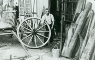 Historische Schwarzweiß-Aufnahme um 1935: Der Stellmacher Heinrich Bosbach vor seiner Werkstatt. Er präsentiert ein selbstgefertigtes hölzernes Wagenrad, das so groß ist wie er selbst.
